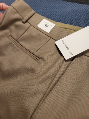 Beige kostymbyxa These Glory Days 46 S - Snygga beige kostymbyxor från These Glory Days i storlek 46. Byxorna har pressveck, smal passform och diskreta fickor. Perfekta för en clean och stilren look. Materialet känns mjukt och har en lätt glansig finish.