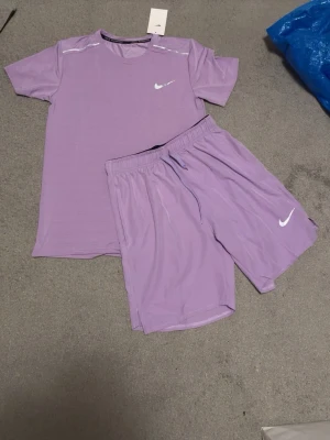 Lila träningsset från Nike - Säljer ett lila träningsset från Nike med t-shirt och shorts. Båda plaggen har Nike-logga i vitt och är tillverkade i ett lätt och snabbtorkande material. Perfekt för gymmet eller löprundan. Skriv för mer info