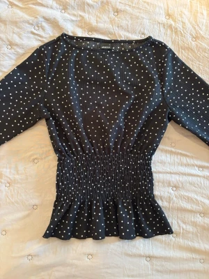 Svart prickig blus från Gina Tricot - Supersöt svart blus från Gina Tricot med vita prickar och smockad midja som ger en snygg siluett. Blusen har lång ärm och lätt rynkad nederkant. Perfekt till jeans eller kjol för en trendig look.