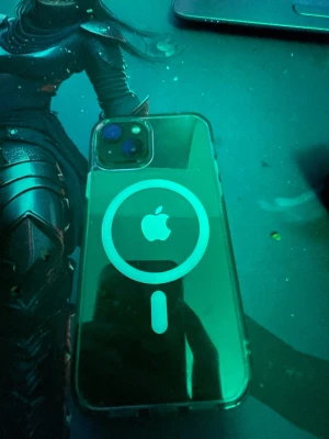 iPhone med MagSafe-kompatibelt skal - Säljer en iPhone med svart framsida och dubbla kameror på baksidan. Telefonen har ett genomskinligt MagSafe-kompatibelt skal med vit cirkel och Apple-logga. Klassisk Apple-design i glas och metall, perfekt för dig som gillar stilren teknik.