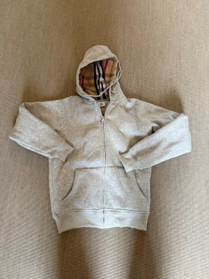 Grå hoodie från Burberry med dragkedja - Säljer en grå hoodie från Burberry med klassiskt rutigt foder i huvan. Tröjan har dragkedja framtill, två stora fickor och ribbade muddar. Materialet är mjukt och bekvämt, perfekt för en chill look. Ikoniskt Burberry-märke i nacken och snygg design på huvan.