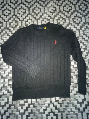 Svart kabelstickad tröja Polo Ralph Lauren - Kabelstickad svart tröja från Polo Ralph Lauren med rund halsringning och röd broderad logga på bröstet. Långa ärmar och ribbade muddar vid ärmslut och nederkant. Klassisk och stilren design som passar till mycket.