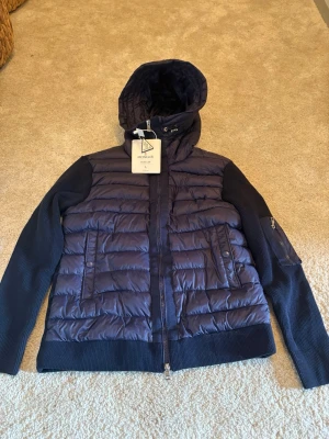 Mörkblå pufferjacka från Moncler - Snygg mörkblå pufferjacka från Moncler med stickade ärmar och sidor, quiltad front och rygg samt huva. Jackan har dragkedja framtill, två fickor med knapp och en ärmficka med Moncler-logga. 