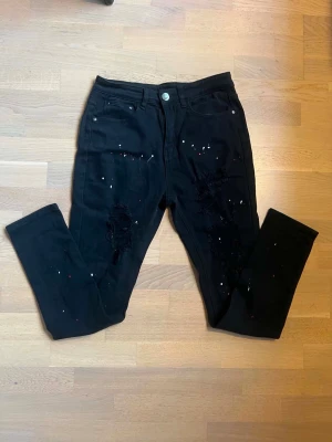 Svarta slitna jeans med färgstänk - Säljer ett par svarta jeans med skinny fit och slitningar på båda benen. Jeansen har coola färgstänk och klassisk femficksmodell. Perfekta för dig som gillar en edgy och trendig stil.