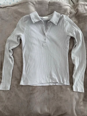 Ljusgrå ribbad långärmad tröja - Säljer en ljusgrå ribbad långärmad polotopp med krage och knappar framtill. Toppen har en slim passform och är tillverkad i mjuk bomullsmix som känns skön mot huden. 