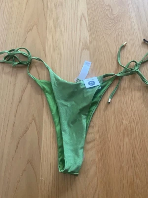 Gröna bikiniunderdel med knytband - Snygg grön bikiniunderdel med smala knytband i sidorna och guldfärgade detaljer på snörena. Tillverkad i glansigt material som ger en lyxig känsla. Perfekt för strand eller poolhäng.