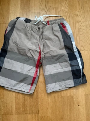 Rutig shorts med snörning - Snygga rutiga shorts i beige, svart, grått och rött med breda ränder och snörning i midjan. Shortsen har elastisk midja och är tillverkade i ett lätt syntetmaterial, perfekta för varma dagar.