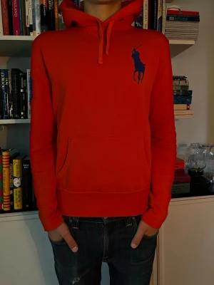 Röd hoodie från Polo Ralph Lauren - Modellen är 179cm väger 65kg. Snygg röd hoodie från Polo Ralph Lauren med stor blå logga på bröstet och en blå siffra 3 på ärmen. Klassisk känguruficka framtill och justerbar huva med dragsko. Perfekt för en avslappnad och sportig stil.