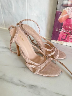 Beige flätade sandaletter från DeeZee - Säljer ett par snygga beige sandaletter från DeeZee med höga klackar och flätade remmar. Skorna har öppen tå och långa snören som lindas runt vristen för en trendig look. Materialet är mockaimitation som ger en mjuk känsla.