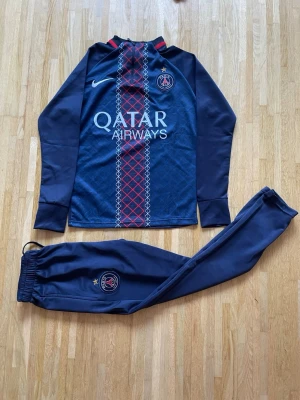 PSG träningsset navy Nike - Snyggt träningsset från PSG i mörkblått med långärmad tröja och matchande byxor. Tröjan har PSG-logga, Nike-logga och Qatar Airways-tryck samt röd och vit detalj framtill. Byxorna har PSG-emblem och resår i midjan. Perfekt för fotbollsträning eller chill.