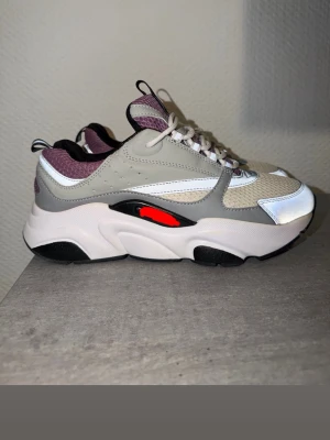 Chunky sneakers i beige och lila - Chunky sneakers med beige mesh och gråa samt vita detaljer. Skorna har lila textilpartier, svarta och röda inslag på sulan samt vita skosnören. Tjock sula i vitt och svart ger en modern look. Perfekta för dig som gillar streetwear och statement-skor.