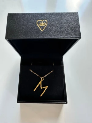 Maria Nilsdotter Wonky letter M necklace  - Säljer ett Wonky letter M gulpläterat silver från Maria Nilsdotter. Kedjans längd är 50cm. Halsbandet är endast använt några få enstaka gånger och skicket har inget märkbart slitage. Box ingår.