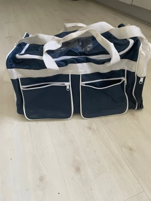 Blå och vit sportig resväska Enda - Säljer en blå och vit sportig resväska från Enda. Väskan har två stora ytterfickor med dragkedja framtill, vita detaljer och breda bärhandtag i tyg. Perfekt för resor eller träning när du vill ha plats för allt du behöver.  Använd 1 gång .