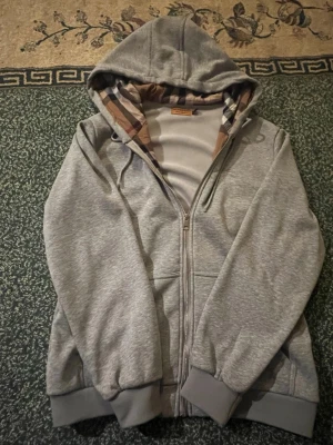 Grå hoodie från Burberry med dragkedja - Snygg grå hoodie från Burberry med klassiskt rutigt mönster i huvan och dragkedja framtill. Hoodien har ribbade muddar och fickor på sidorna. Perfekt för en chill och stilren look.