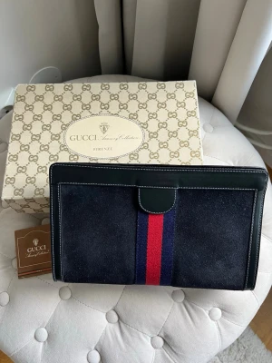 Gucci väska / clutch / kuvertväska  - Supersnygg vintage Gucci clutch i blått med röd och blå rand. Utanpå är väskan som ny, aningen sliten inuti men inget som påverkar väskans utseende.  Väskan har en invändig etikett med ”GUCCI Accessory Collection MADE IN ITALY” och ett serienummer ”67-014-2126”. Den kommer med original Gucci-låda och ett litet häfte. Mått ca: 27.5 x 17 x 8 cm. 100% äkta.  Katt finns i hemmet.  Kan hämtas eller skickas.