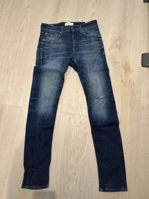 Blå slim fit jeans från Calvin Klein Jeans - Säljer ett par blå slim fit jeans från CK Jeans med snygg slitning och en liten reva på ena benet. Jeansen har klassisk femficksmodell, skinnpatch bak i midjan och är tillverkade i stretchig denim för skön passform. Sparsamt använda. Jag är 183 cm lång och bär S/M. W29 L32. 