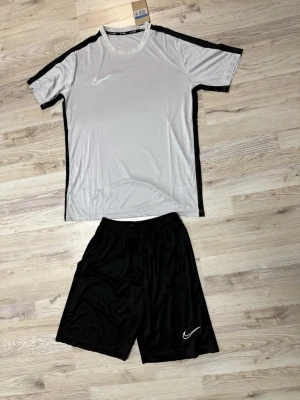 Nike träningsset svart och vitt - Snyggt träningsset från Nike med en vit t-shirt med svarta detaljer längs sidor och ärmar samt ett par svarta shorts. Båda plaggen har Nike-loggan tryckt framtill. Perfekt för gymmet eller fotbollsträningen. Tillverkade i ett lätt och snabbtorkande material.