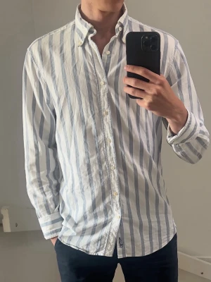 Vit och blå randig skjorta Stenströms - Klassisk vit skjorta med blåa vertikala ränder och knappar framtill. Skjortan har lång ärm, button-down krage och är tillverkad i ett lätt bomullstyg. Perfekt för en clean och fräsch stil.