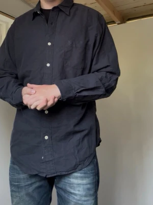 Vintage Ralph Lauren | Skjorta | L - Klassisk svart skjorta från Polo Ralph Lauren med vit knappstängning och bröstficka. Skjortan har lång ärm och är tillverkad i bomull, perfekt för en stilren look. Mått: Längd 73 Bredd 58