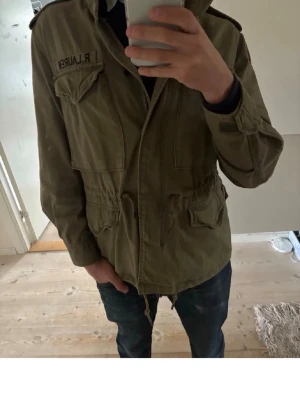 Field jacket från Ralph Lauren - Snygg Field jacket från Ralph Lauren med patchad amerikansk flagga på ärmen och snörning i midjan. Jackan har flera fickor framtill, axelklaffar och dragkedja. Perfekt för dig som gillar utility-stil och vill ha en cool vardagslook.