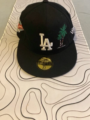 Svart LA New Era 59FIFTY keps - Snygg svart keps från New Era 59FIFTY med LA-logga framtill, broderade palmer och World Series-märke på sidan. Rak skärm och klassisk passform. Perfekt för dig som gillar streetwear och sportig stil.