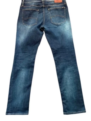 Tommy Hilfiger Jeans - Snygga blå jeans från Tommy Hilfiger med raka ben och klassisk femficksmodell. Jeansen har slitningar och tvättade detaljer för en cool look. Tillverkade i denim med en skön passform och läderpatch bak i midjan.