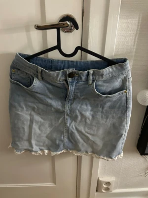 Ljusblå jeanskjol med spetskant - Säljer en ljusblå jeanskjol med spetskant längst ner. Kjolen har klassisk femficksdesign, bälteshällor och knappstängning fram. Perfekt för dig som gillar en söt och avslappnad stil.