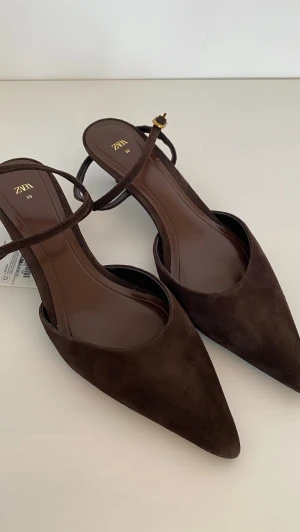 Bruna mockapumps från Zara - Snygga bruna pumps från Zara i mocka med spetsig tå och smal rem med guldfärgat spänne runt ankeln. Skorna har en elegant och stilren design som passar perfekt till många olika outfits. Klackhöjden är låg till medelhög och materialet känns lyxigt.