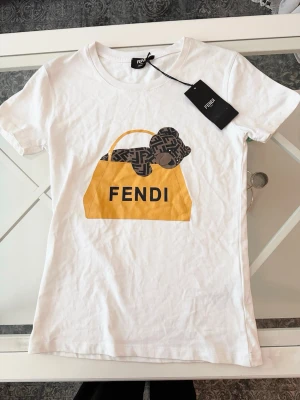 Vit Fendi t-shirt med nalletryck - Vit t-shirt med ett stort tryck av en gul väska och en brun nalle med mönster på bröstet. Klassisk rund hals och korta ärmar. Perfekt för dig som gillar exklusiva och lekfulla plagg.