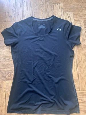 Svart träningstshirt från Under Armour - Snygg svart t-shirt från Under Armour med diskret logga på bröstet. T-shirten har korta ärmar och är tillverkad i ett mjukt och stretchigt syntetmaterial som andas, perfekt för träning. Figurnära passform och klassisk rund halsringning.