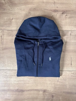 Mörkblå hoodie från Polo Ralph Lauren - Snygg mörkblå hoodie från Polo Ralph Lauren med dragkedja framtill och klassisk huva. Ikoniska vita polospelar-loggan broderad på bröstet. Tillverkad i mjukt bomullsmaterial och har dragsko i huvan för justerbar passform.
