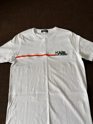 Vit t-shirt från Karl Lagerfeld - ALDRIG ANVÄNT DÅ DEN VSR FÖR LITEN!!Snygg vit t-shirt från Karl Lagerfeld med en röd linje över bröstet och logga tryckt på vänster sida. Klassisk rund hals och korta ärmar. Tillverkad i mjuk bomull som känns skön mot huden. Perfekt för en clean och stilren look.