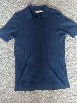 Marinblå ribbstickad polo från Kappahl - Snygg marinblå polotopp från Kappahl med ribbad struktur och korta ärmar. Klassisk krage med knappslå framtill ger en stilren look. Perfekt till jeans eller kjol för en trendig outfit.