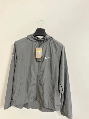 Nike Miller  - Snygg grå vindjacka från Nike med dragkedja framtill och justerbar huva. Jackan har en minimalistisk design med vit Nike-logga på bröstet och praktiska sidofickor. Tillverkad i lätt och vindtätt material, perfekt för blåsiga dagar.
