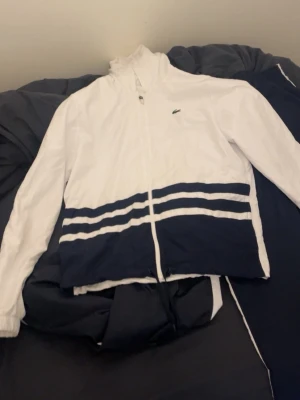 Lacoste träningsset i vitt och marinblått - Snyggt träningsset från Lacoste med vit zip-jacka och marinblå byxor. Jackan har marinblå detaljer och vita ränder nertill samt dragkedja och hög krage. Byxorna är helmarinblå med diskret Lacoste-logga och vita detaljer längs sidorna. Perfekt för sport eller chill.