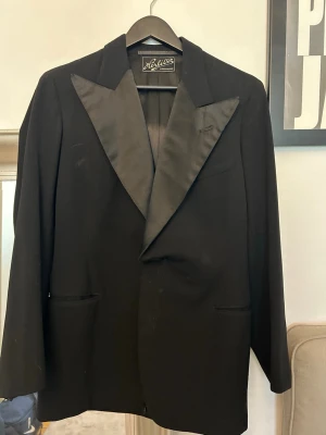 Svart vintage kavaj - Stilren svart kavaj från Hörlits i Landskrona. Kavajen har breda slag i satin och klassisk passform, samt helfodrad insida i blankt material. Perfekt för dig som vill ha en elegant look till speciella tillfällen. Kavajen är i storlek M/L och är lite längre än en vanlig kavaj