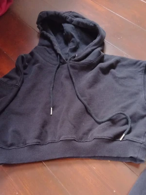 Svart croppad hoodie med huva - Säljer en svart croppad hoodie med huva och dragsnören. Hoodien har ribbade muddar nedtill och är tillverkad i mjukt sweatshirtmaterial. Perfekt för en avslappnad och trendig look.