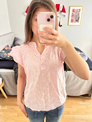 Ljusrosa blus med broderade blommor - Söt ljusrosa blus med broderade blommor och korta ärmar. Den är tillverkad i ett tunt och luftigt tyg.