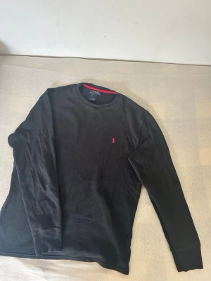 Svart långärmad topp Polo Ralph Lauren - Klassisk svart långärmad topp från Polo Ralph Lauren i 100% bomull. Tröjan har rund halsringning, diskret röd logga broderad på bröstet och en snygg röd detalj på insidan av kragen. Perfekt för en stilren och enkel look.