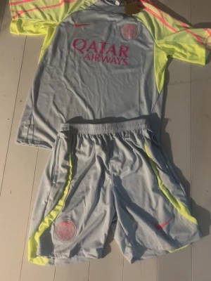 PSG träningsset grå/gul Nike - Säljer ett PSG träningsset från Nike i ljusgrått med neon-gula och rosa detaljer. Tröjan har PSG-logga och Qatar Airways-tryck på bröstet, samt matchande shorts med elastisk midja. Materialet är lätt och andas, perfekt för fotbollsträning.