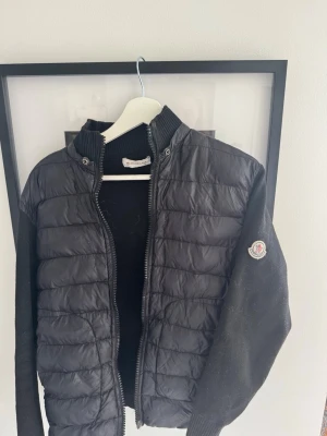 Svart cardigan från Moncler - Snygg svart dunjacka från Moncler med quiltad front och stickade ärmar. Jackan har hög krage, dragkedja och Moncler-logga på ena ärmen. Perfekt för kalla vinterdagar och ger en stilren look.