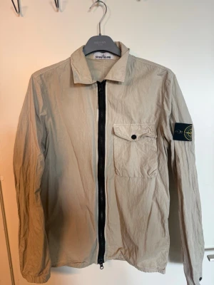 Beige overshirt från Stone Island - Stilren beige overshirt från Stone Island med svart dragkedja framtill, bröstficka med knapp och ikonisk logotyp-patch på vänster ärm. Perfekt nu inför sommaren! Jackan är i väldigt gott skick, den har smuts/fläckar vid kragen (se bild), något som bör gå bort i tvätten. Utöver det har den inga skador eller defekter, den kan behövas strykas då den inte använts på flera år.