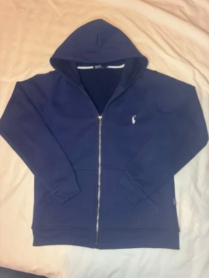Marinblå hoodie från Polo Ralph Lauren - Snygg marinblå hoodie med dragkedja från Polo Ralph Lauren. Klassisk design med vit broderad logga på bröstet, huva och ribbade muddar. Perfekt för en chill och stilren look. Tillverkad i mjukt bomullsmaterial. Knappt använd. Priset kan diskuteras snabb frakt!