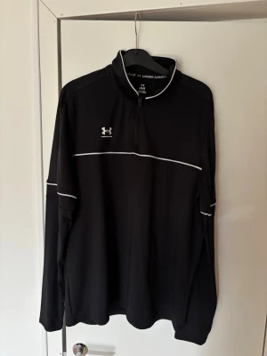 Svart långärmad tröja Under Armour - Svart långärmad sporttröja från Under Armour med vit piping längs bröstet och ärmarna. Tröjan har halv dragkedja och hög krage, perfekt för träning eller löpning. Materialet är syntetiskt och känns lätt och smidigt.