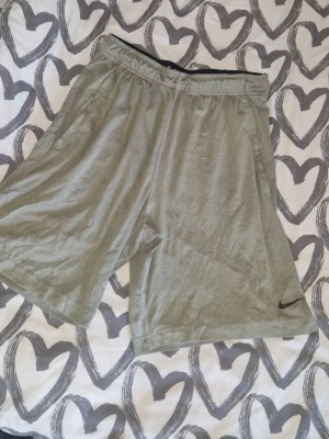 Nike shorts - Säljer ett par grå shorts från Nike med elastisk midja och klassisk svart swoosh-logga på benet. De är tillverkade i mjukt bomullsmaterial och har en loose passform som är perfekt för träning eller chill dagar. Väl använda men inga defekter förutom ett litet hål och att loggan har någon tråd-se bilder.