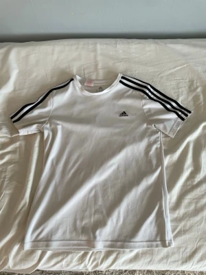 Adidas T-Shirts - (Pris kan diskuteras) Säljer en vit Adidas t-shirt med klassiska svarta ränder på axlarna och logga på bröstet. T-shirten är kortärmad och gjord i ett mjukt och lätt material, perfekt för sport eller chill. Enkel och stilren design som funkar till det mesta.