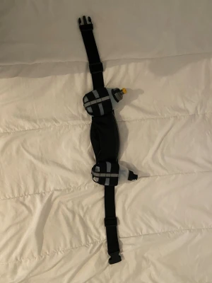Svart löparbälte med flaskhållare - Praktiskt svart löparbälte med justerbart spänne och två små flaskhållare med reflexdetaljer. Perfekt för att bära småsaker och vattenflaskor under träning eller promenad. Tillverkad i slitstarkt syntetmaterial och har smarta fack för enkel förvaring.