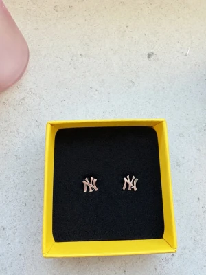 New Era örhängen  - Säljer ett par snygga örhängen i form av New York Yankees-loggan. De är i roséguldfärgad metall och har en stilren, modern look. Perfekta för dig som gillar sportiga accessoarer med en touch av streetstyle.