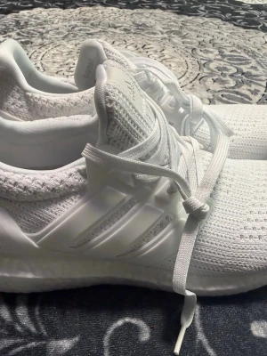 Vita Adidas Ultraboost sneakers - Säljer ett par helvita Adidas Ultraboost sneakers med stickad ovandel och Boost-sula för extra komfort. Skorna har klassiska tre ränder på sidan, vita skosnören och en låg silhuett. Perfekta för dig som gillar stilrena och sportiga sneakers.