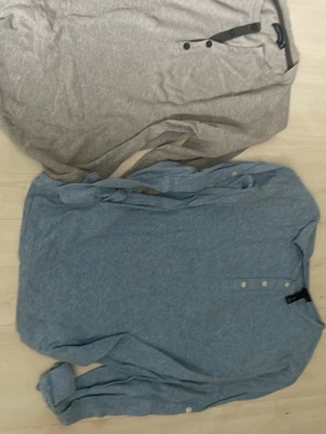 Två långärmade tröjor med knapparPoco Loco - Säljer två långärmade tröjor, en ljusgrå och en ljusblå. Båda har knappdetaljer vid halsen och är tillverkade i mjukt, tunt material. Perfekta för en avslappnad stil och passar bra till jeans eller chinos.